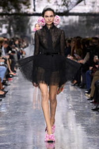 Model walking a runway in a black lace dress with a voluminous skirt, pink flower hair accessories, and pink floral shoes.