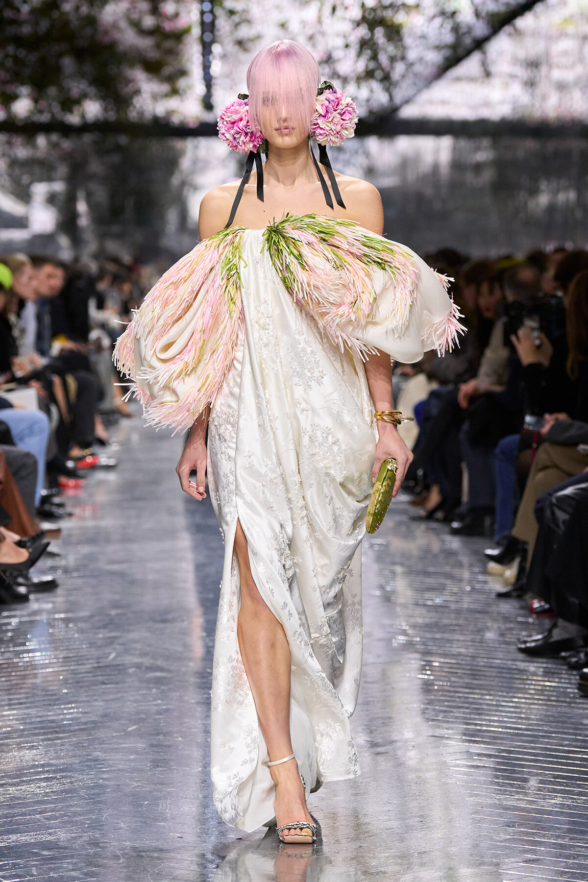 Model on a runway wearing a voluminous white embroidered gown with pink feathered sleeves and a pink floral headpiece surrounding her short pink hair, walking the catwalk.”