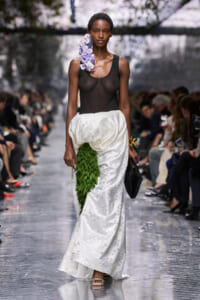 Model walks runway wearing a sheer black top and a white embroidered skirt with a green leaf appliqué, purple floral accessory at the shoulder, and sandals.