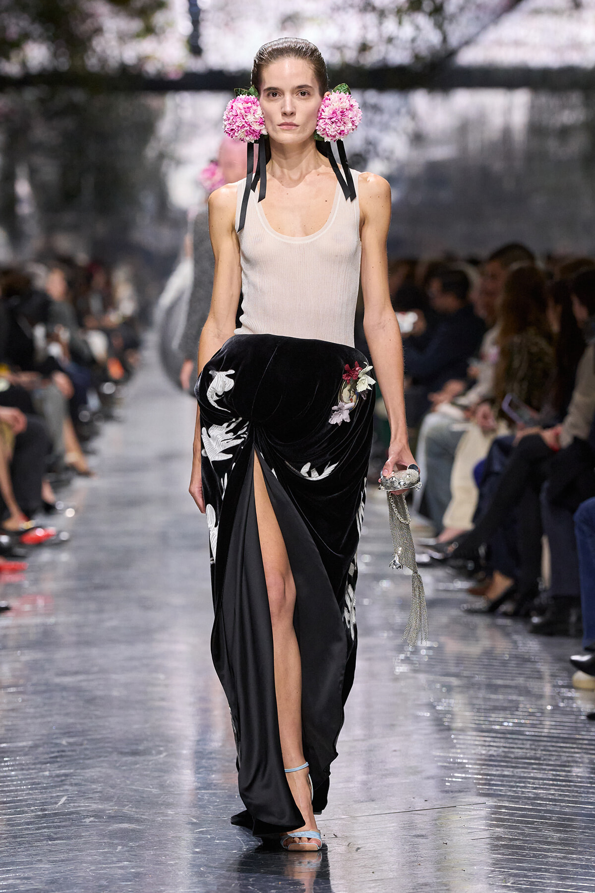 Model walks runway in a beige sleeveless top and black velvet skirt with a high slit and white floral embroidery, pink flower headpieces, holding a silver tasseled clutch.