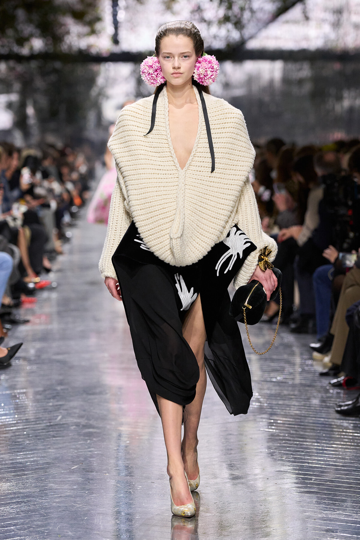 Runway model in an oversized cream knit sweater, black skirt with white bird print, pink floral ear accessories, and beige heels.