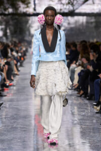 Model walking the runway in a pale blue blazer with black lapels, a layered white feathered skirt, and pink floral headpieces.