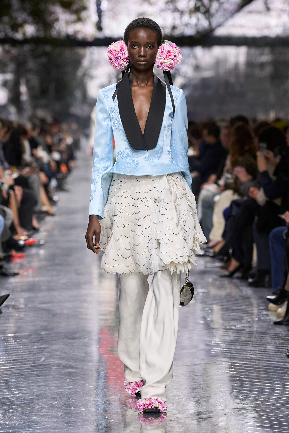 Model walking the runway in a pale blue blazer with black lapels, a layered white feathered skirt, and pink floral headpieces.