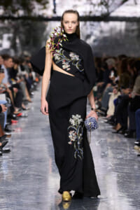 Model walking runway in a black embroidered gown with floral motifs and a cape, holding a blue patterned clutch and wearing gold shoes.