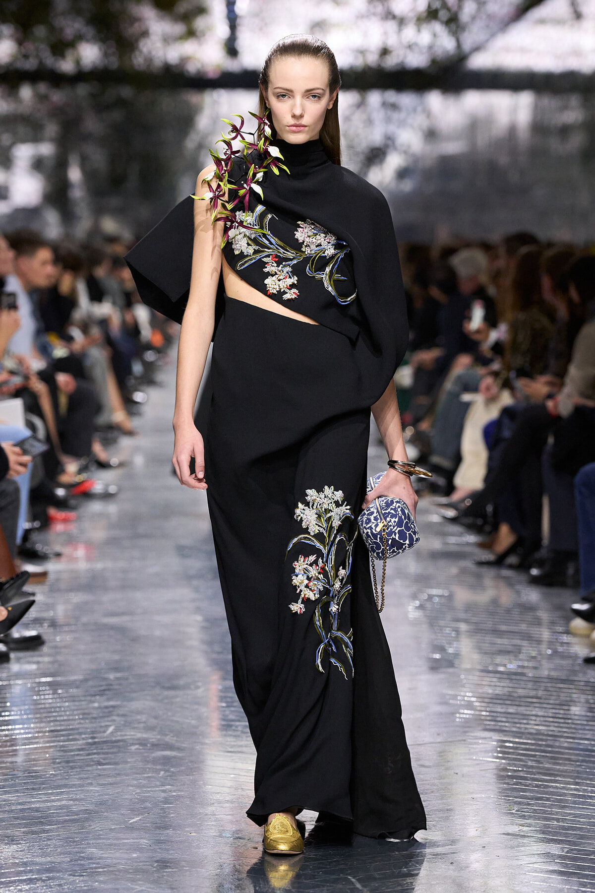 Model walking runway in a black embroidered gown with floral motifs and a cape, holding a blue patterned clutch and wearing gold shoes.