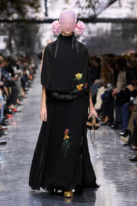 Model walks the runway in a black, floor-length gown with embroidered flowers; oversized sleeves and a pink wig with large flower adornments on both sides, holding a small striped handbag with a gold chain and wearing gold shoes.