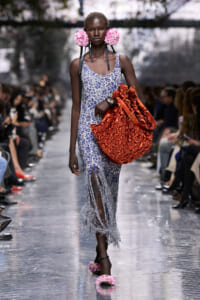 Model on a runway wearing a blue floral dress with fringe hem, pink flower earrings, and a large red ruched handbag.