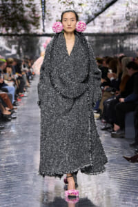 Model walks the runway in a oversized, textured gray tweed coat with fringe, wearing pink flower accessories behind the ears and matching pink fluffy sandals on a reflective runway.