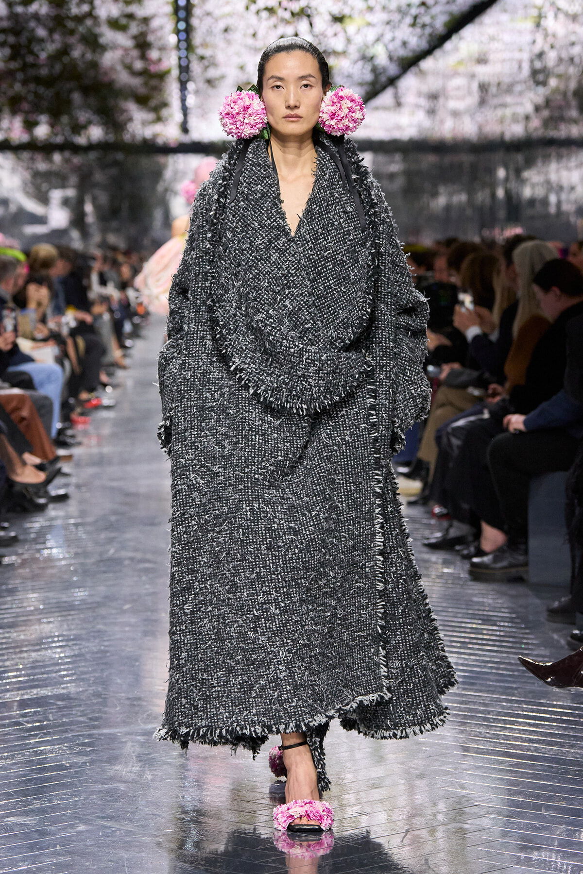 Model walks the runway in a oversized, textured gray tweed coat with fringe, wearing pink flower accessories behind the ears and matching pink fluffy sandals on a reflective runway.