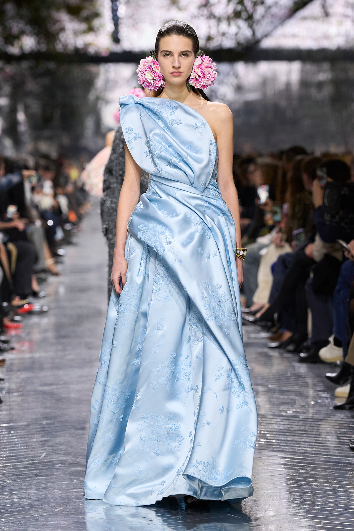 Model on a runway wearing a pale blue one-shoulder satin gown with floral embroidery; pink flowers in her hair.