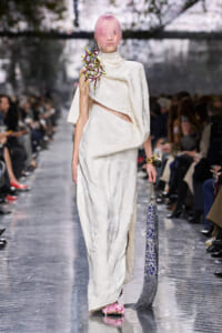 Runway model wears an ivory floral gown with cape, pink hair, and carries a blue patterned clutch; floral accessory on shoulder.