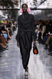 Model walks a fashion runway in a dark knit sweater and wide black trousers, with pink flower earrings and a small orange handbag en route to the camera.