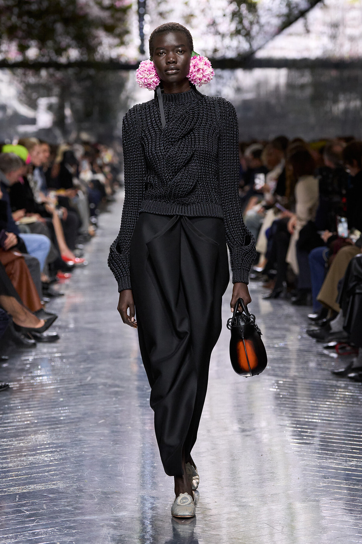 Model walks a fashion runway in a dark knit sweater and wide black trousers, with pink flower earrings and a small orange handbag en route to the camera.