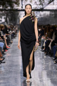 Model walking the runway in a black draped one-shoulder gown with an orchid on the shoulder and a high slit, holding a floral clutch.