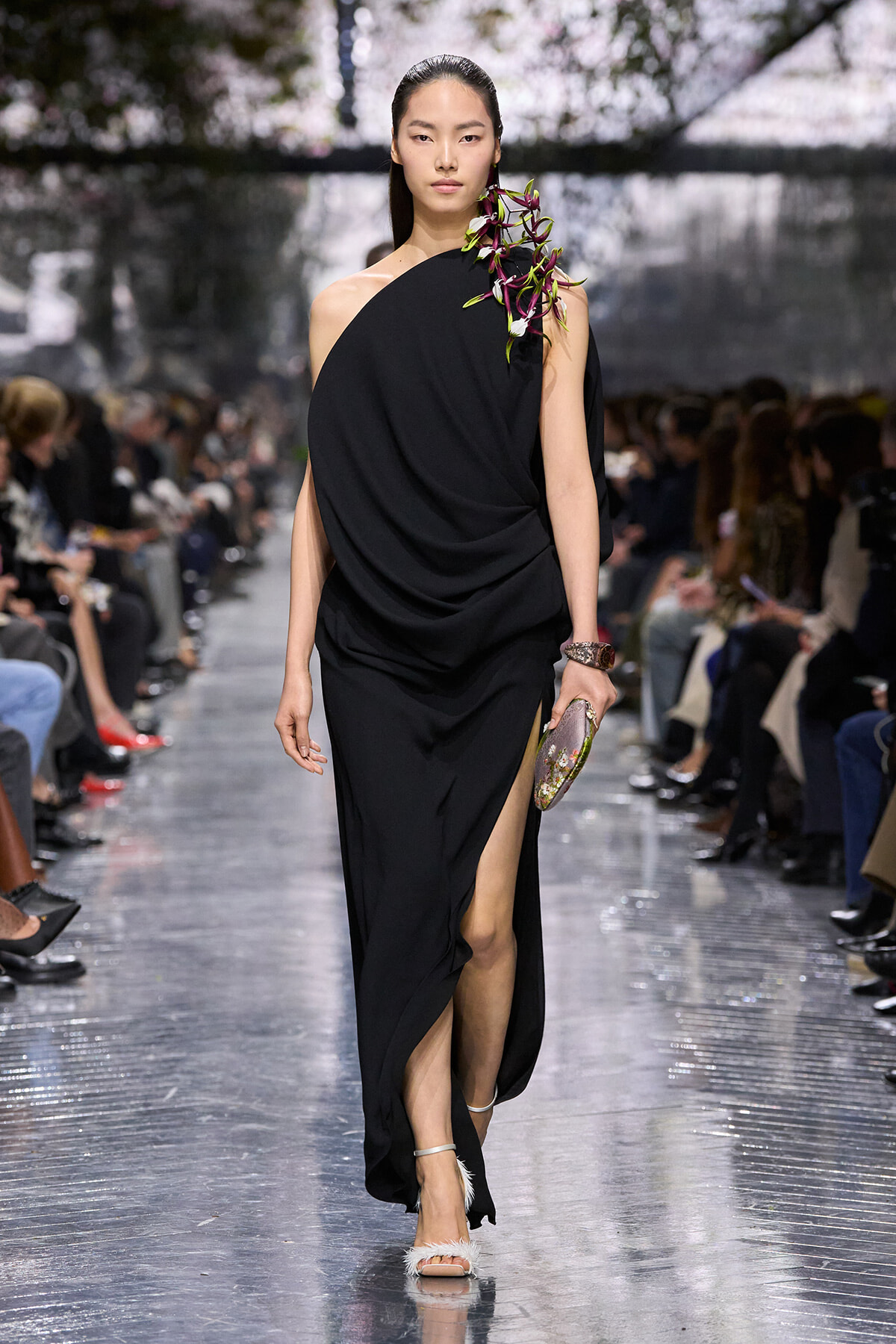 Model walking the runway in a black draped one-shoulder gown with an orchid on the shoulder and a high slit, holding a floral clutch.