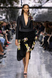 Runway model in a black tailored blazer with a deep V, paired with a black textured skirt adorned with pink and yellow floral clusters, walking the catwalk.