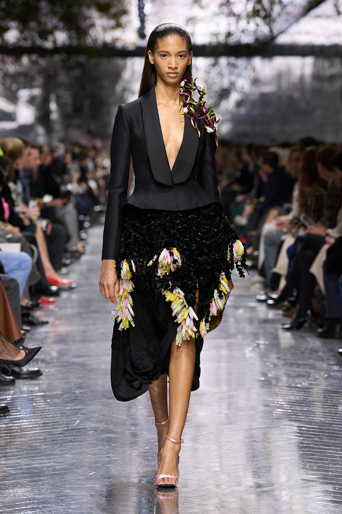 Runway model in a black tailored blazer with a deep V, paired with a black textured skirt adorned with pink and yellow floral clusters, walking the catwalk.