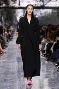 Model walks a runway in a long black V-neck coat, paired with pink fluffy shoes; audience blurred in background.