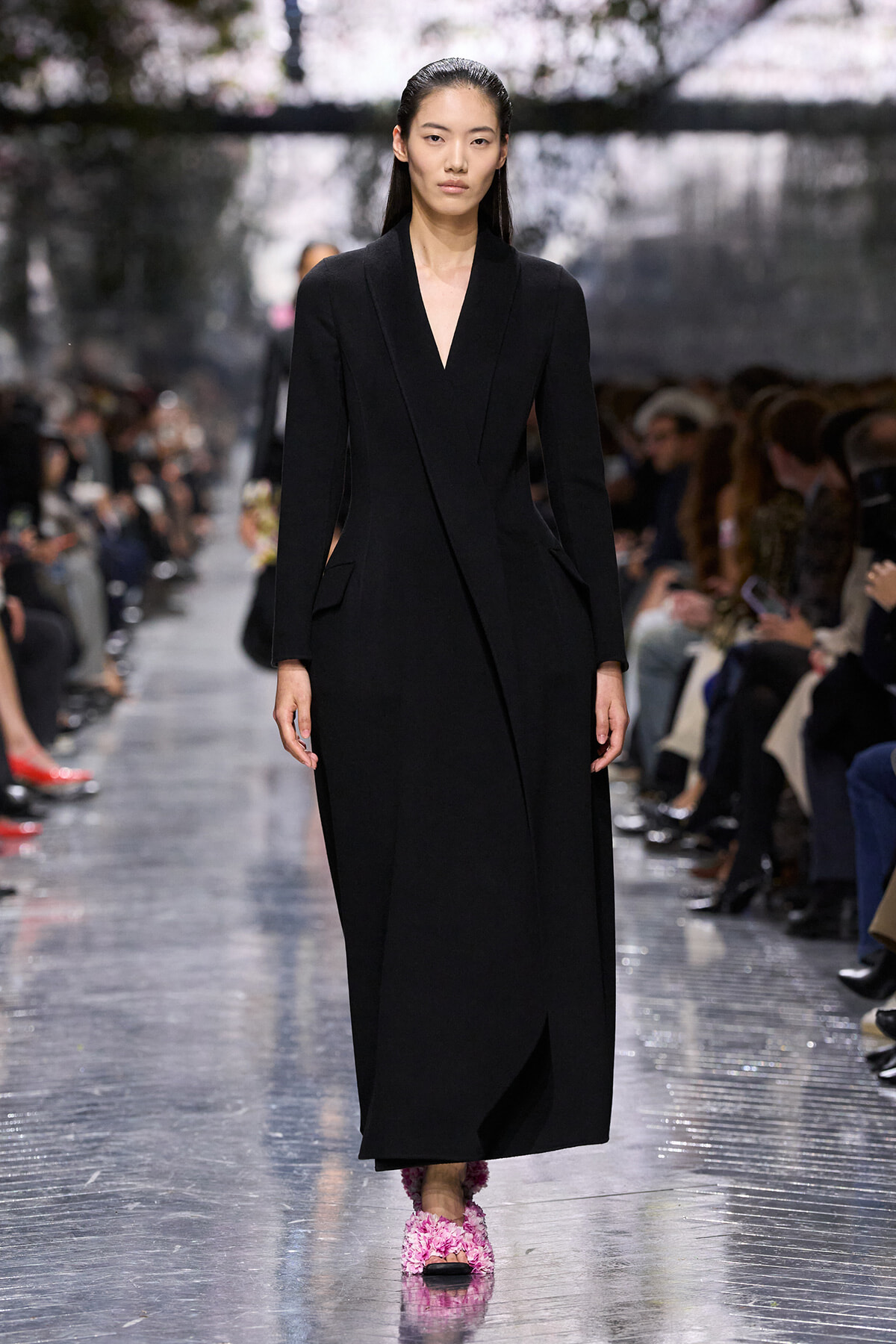 Model walks a runway in a long black V-neck coat, paired with pink fluffy shoes; audience blurred in background.