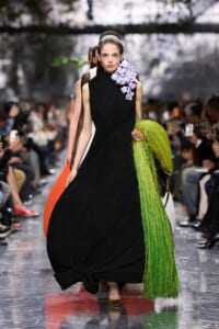 Model walks the runway in a black pleated, floor-length gown with a purple floral accent at the shoulder, holding a vivid lime-green fringe scarf.