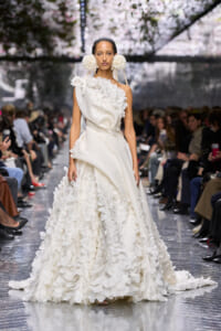 Model wearing a white couture gown with layered petal embellishments on a fashion-show runway, audience in background.