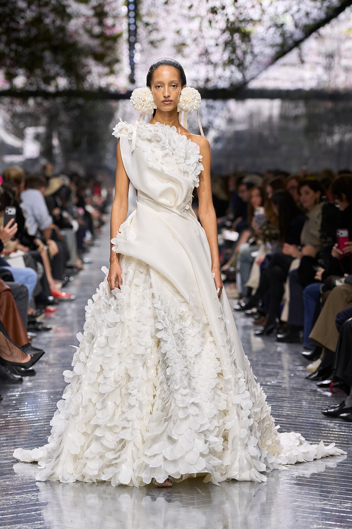 Model wearing a white couture gown with layered petal embellishments on a fashion-show runway, audience in background.