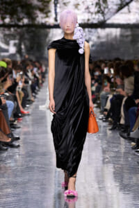 Model walking a runway in a black draped velvet dress with a lavender fringe hairstyle and a purple floral accessory over the shoulder, carrying an orange handbag. Note: keep concise.