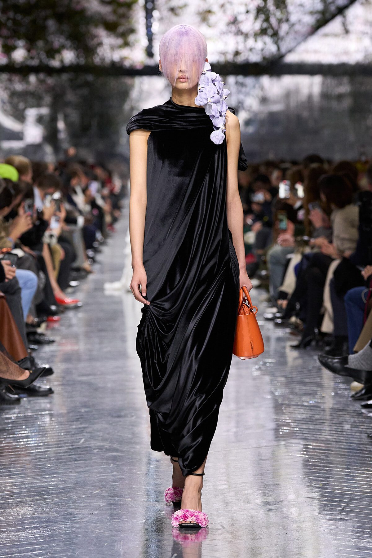 Model walking a runway in a black draped velvet dress with a lavender fringe hairstyle and a purple floral accessory over the shoulder, carrying an orange handbag. Note: keep concise.