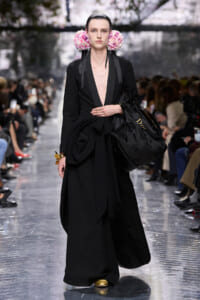 Female model on a fashion-show runway in a long black outfit with a deep V neckline, pink floral ear adornments, and a large black Dior tote with gold shoes beneath.