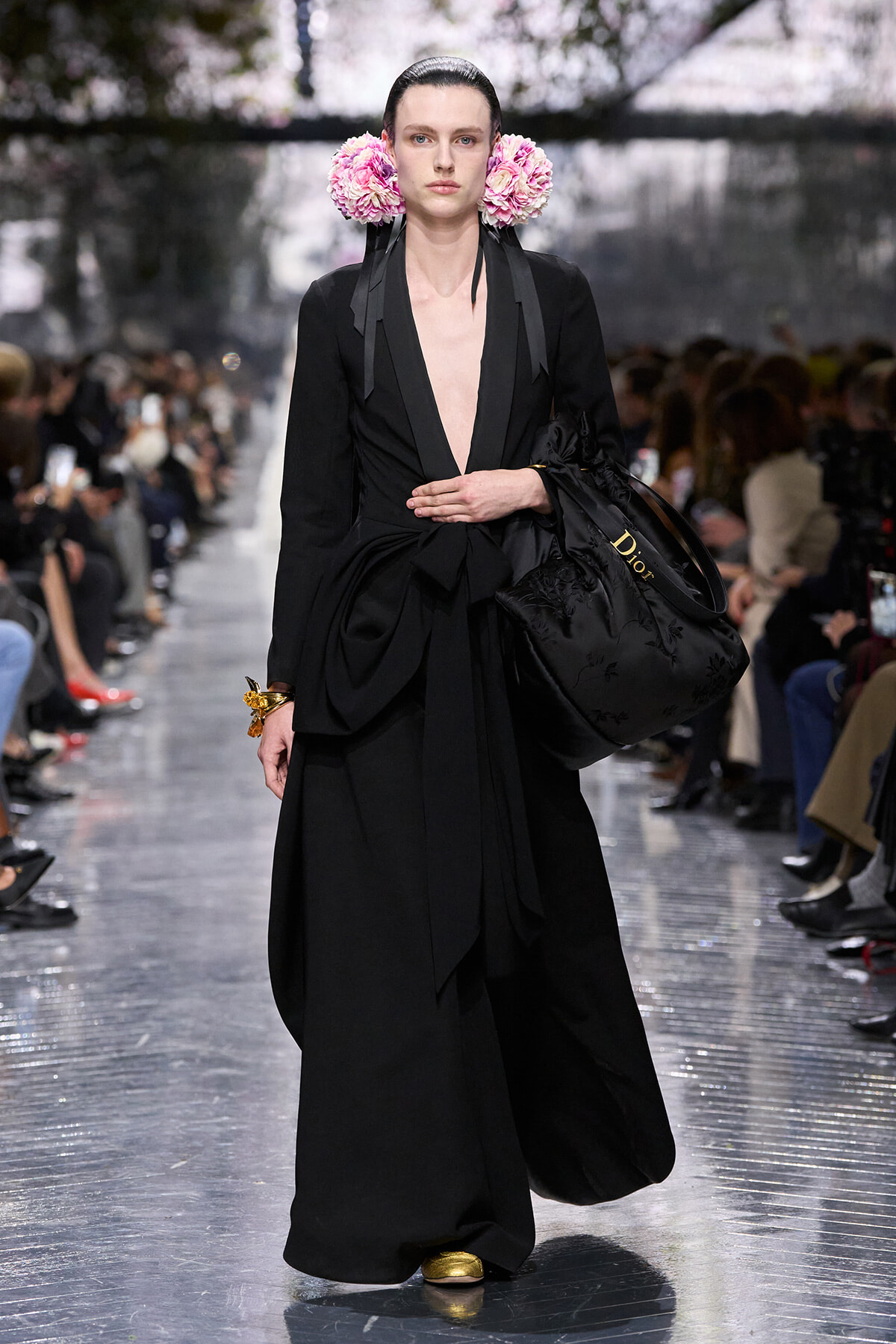 Female model on a fashion-show runway in a long black outfit with a deep V neckline, pink floral ear adornments, and a large black Dior tote with gold shoes beneath.