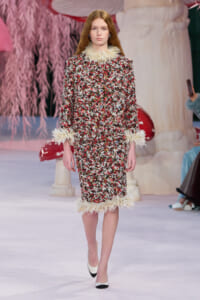 Model walking the runway in a multicolor tweed dress with cream feather trim at the collar, cuffs, and hem.