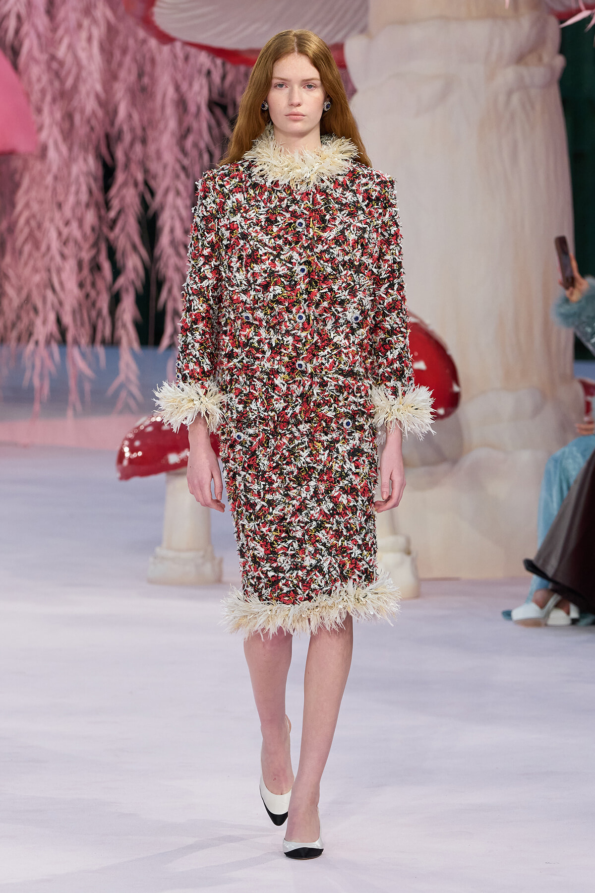 Model walking the runway in a multicolor tweed dress with cream feather trim at the collar, cuffs, and hem.