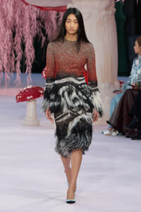 Model walking the runway in a red-to-black gradient knit dress with layered black-and-white feather fringe at the skirt, turquoise heels.