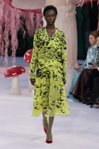 Model on a fashion show runway wearing a neon yellow-green dress with a black abstract pattern and tiered skirt; pink decor in the background.