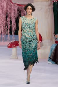 Model walking the runway in a sleeveless teal-to-green crochet dress with leaf-like lace pattern.