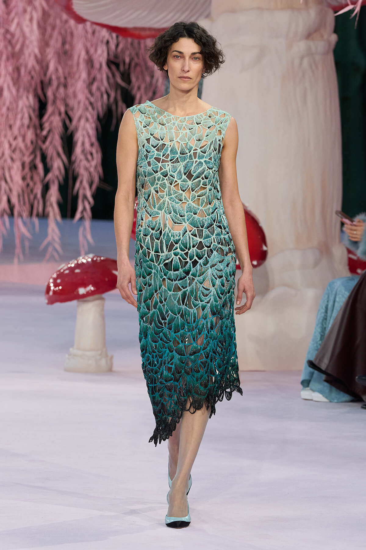 Model walking the runway in a sleeveless teal-to-green crochet dress with leaf-like lace pattern.