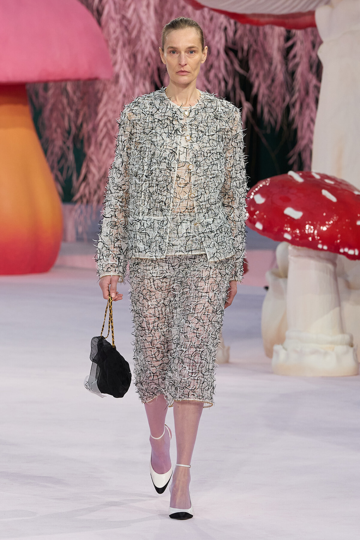 Model walks the runway in a silver textured tweed suit with a sheer underlayer, holding a small black handbag.