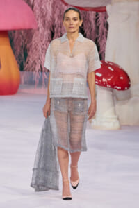 Model walks a runway in a sheer gray two-piece outfit with large front pockets, holding a matching sheer scarf at her side.