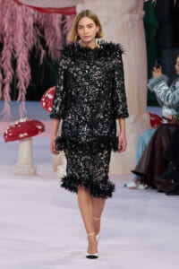 Model on a runway wearing a black lace textured coat with feathered trim and matching knee-length skirt, white heels, and minimal makeup.
