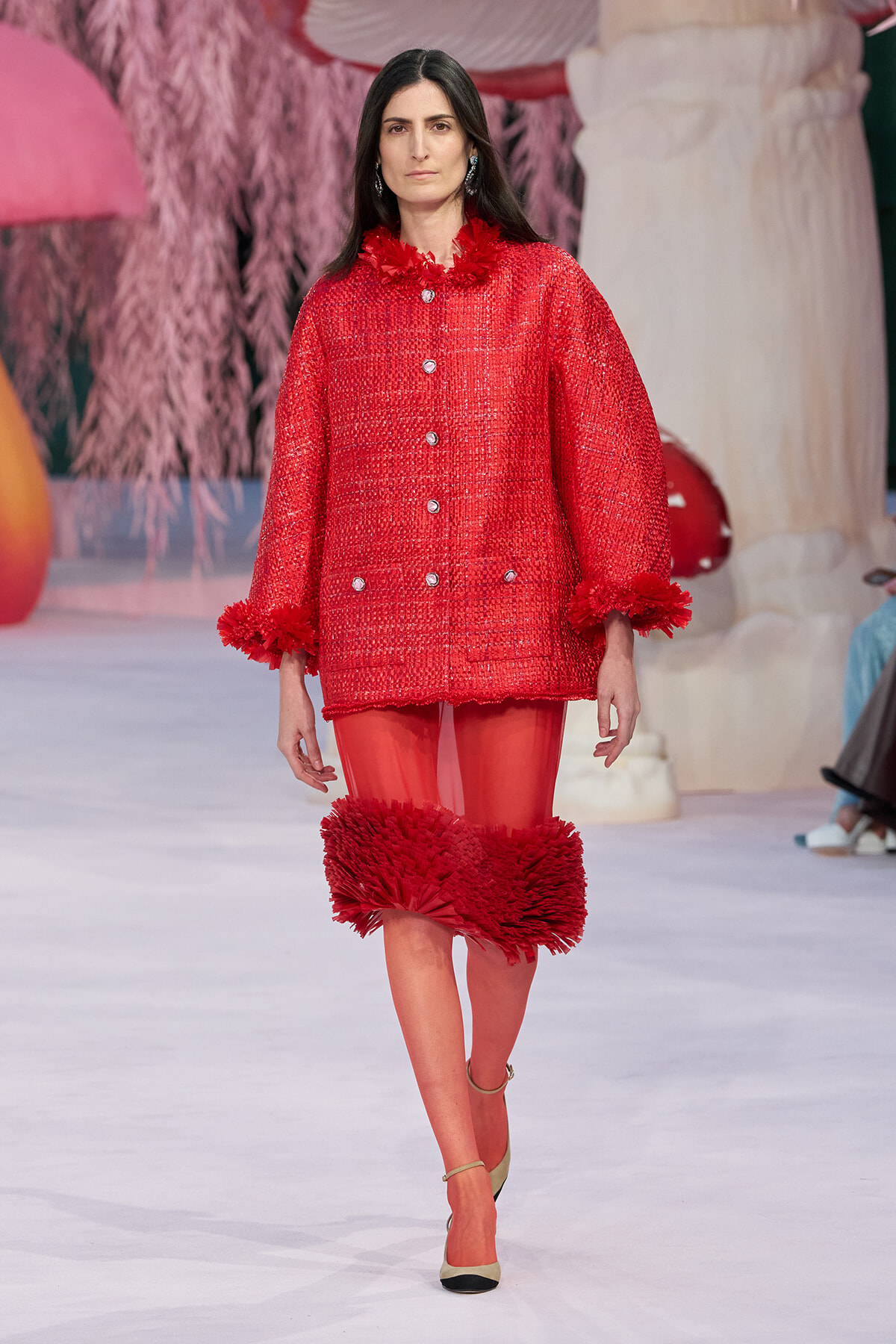 Runway model wearing a red textured coat with feathered cuffs and hem, red tights, and beige pumps.