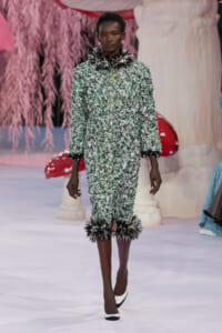 Model walks a runway in a green-and-white textured suit with black feather trim at the cuffs and hem.