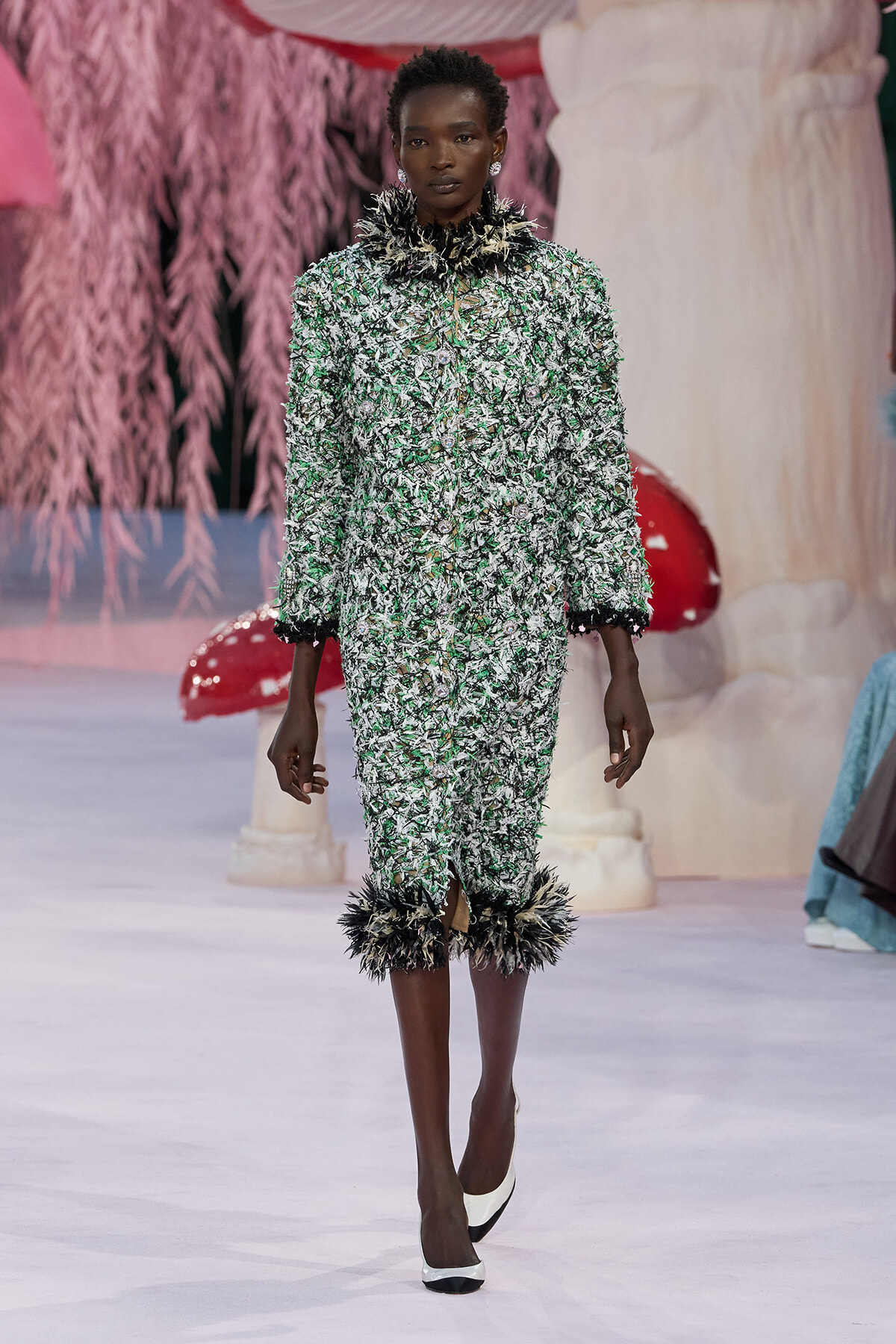 Model walks a runway in a green-and-white textured suit with black feather trim at the cuffs and hem.