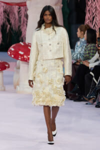 Model walking the runway in a cream textured two-piece suit with floral appliqué detailing, white shoes, and minimal makeup.