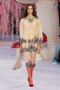 Model walking the runway in a sheer pale pink coat and matching shorts, with bright mushroom-and-floral embroidery at the hem and collar, carrying a beige bag.