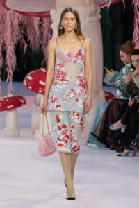 Model walking the runway in a sheer light-blue dress with red and white floral embroidery, carrying a pink quilted handbag and wearing two-tone sandals; audience seated in the background.
