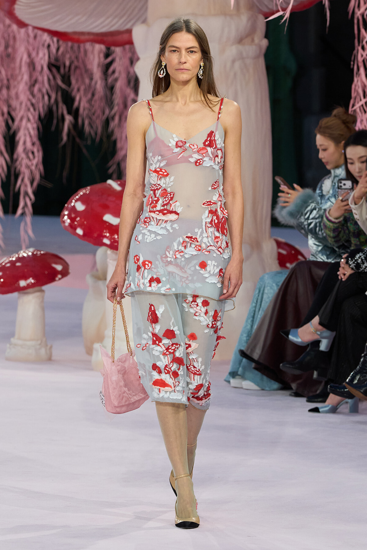 Model walking the runway in a sheer light-blue dress with red and white floral embroidery, carrying a pink quilted handbag and wearing two-tone sandals; audience seated in the background.