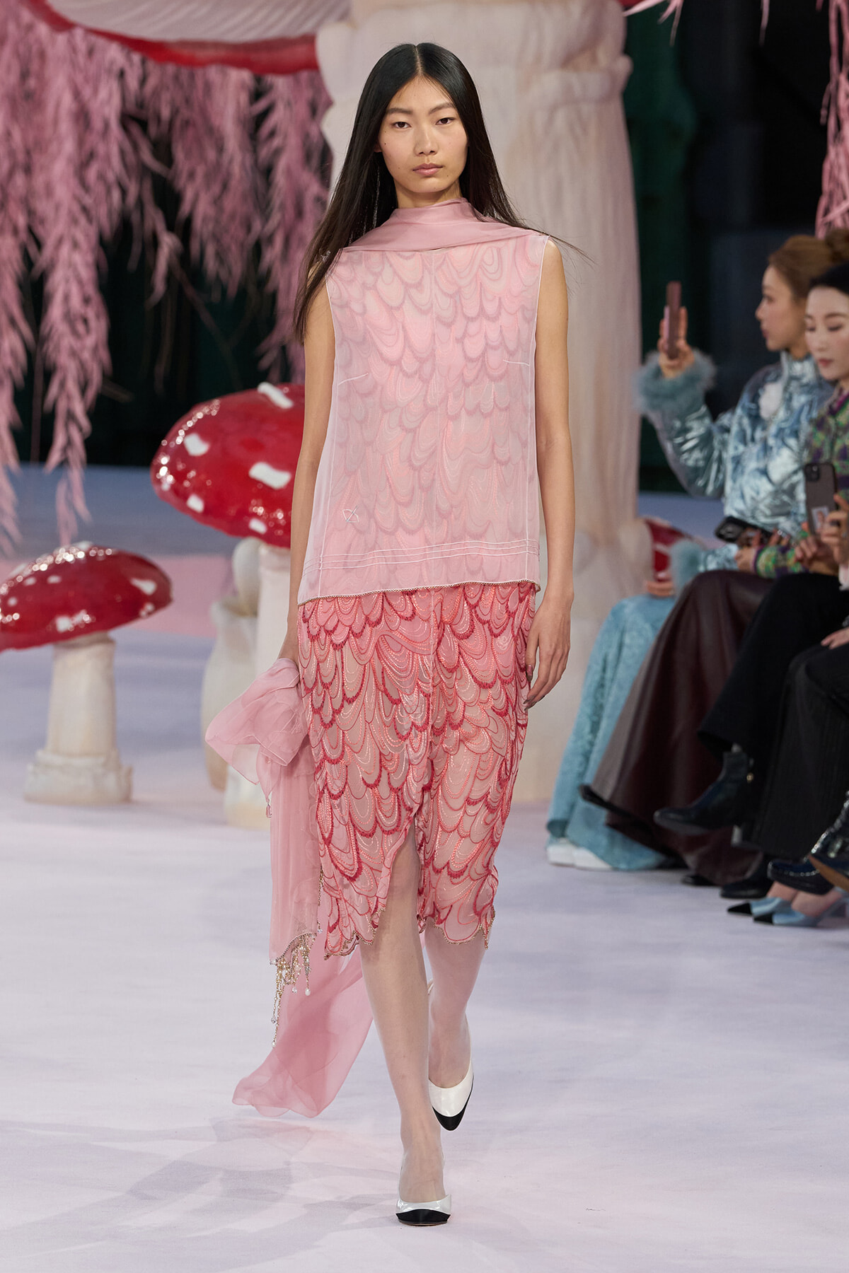 Model walks the runway in a pink layered dress with scalloped lace patterns, holding a sheer pink scarf, facing forward amid a whimsical set with oversized mushrooms.