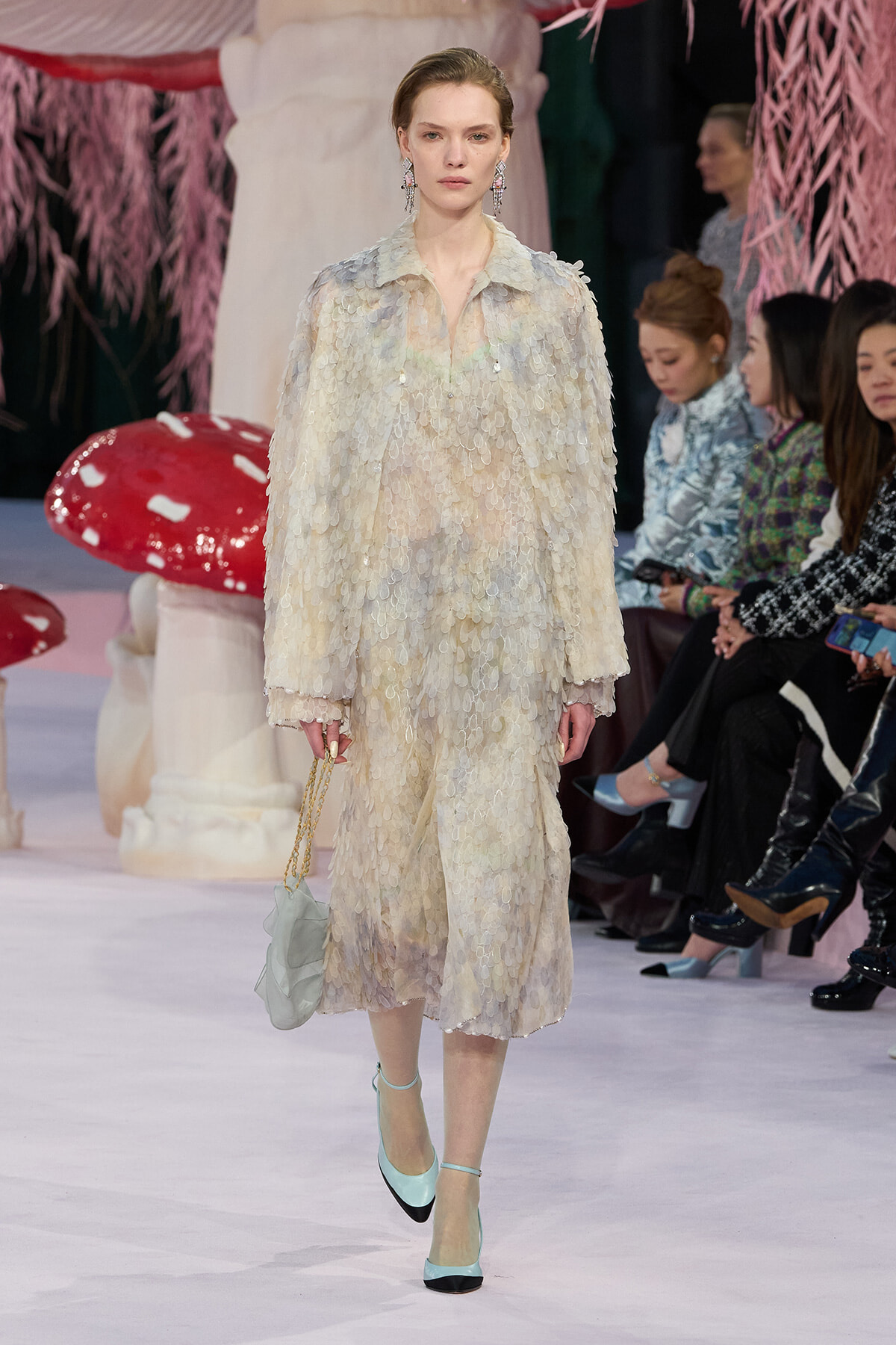 Model walks the runway in a pale, iridescent, scalloped-sequin coat-dress with pastel hues, mint-green shoes, and a small pale handbag.