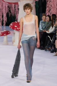 Model on a runway wearing a sheer pale pink tank top and light patchwork denim jeans, holding a gray fringed scarf while spectators watch in the background.