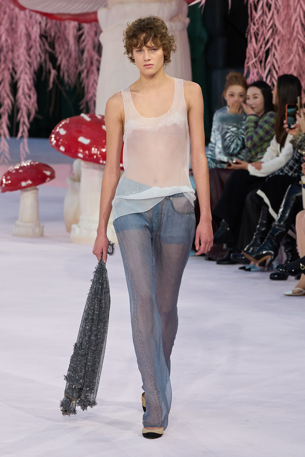 Model on a runway wearing a sheer pale pink tank top and light patchwork denim jeans, holding a gray fringed scarf while spectators watch in the background.